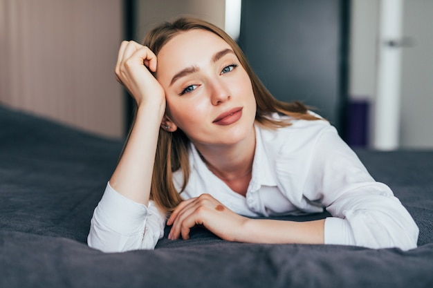 Jonge vrouw liggend aan het einde van het bed onder het dekbed en glimlachend, met haar hoofd op haar hand rustend met de andere in het haar