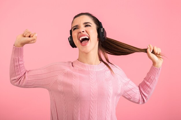 Jonge vrouw luisteren naar muziek in draadloze koptelefoon dragen roze trui glimlachend poseren op roze