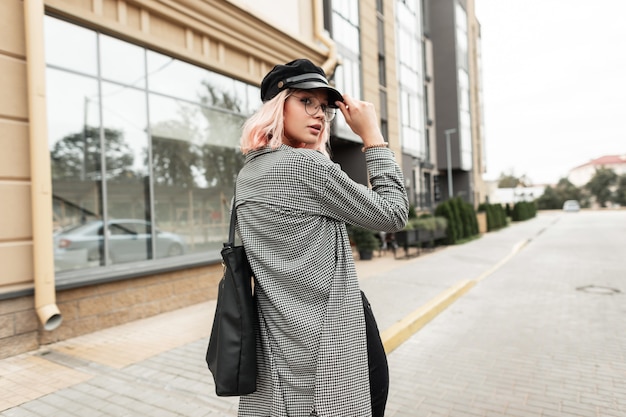 Jonge vrouw mannequin in stijlvolle bril met een vintage hoed in een modieus geruit hemd met een tas loopt op straat in de buurt van het gebouw