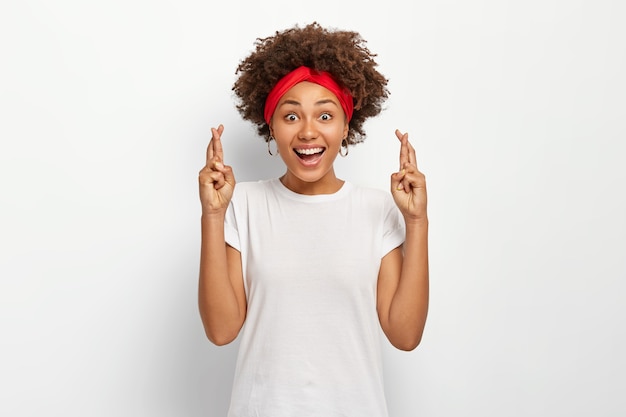 Jonge vrouw met Afro-kapsel, kruist vingers voor geluk, wacht op wonderen, draagt rode hoofdband en casual t-shirt