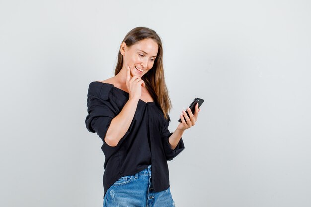 Jonge vrouw met behulp van smartphone in shirt, korte broek en op zoek vrolijk. vooraanzicht.