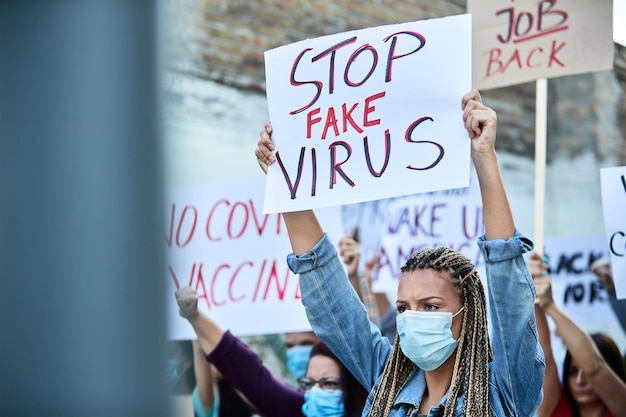 Jonge vrouw met beschermend gezichtsmasker met banner met 'stop nep virus' inscriptie terwijl ze protesteert met een menigte mensen tegen afsluitingen als gevolg van een coronavirusepidemie