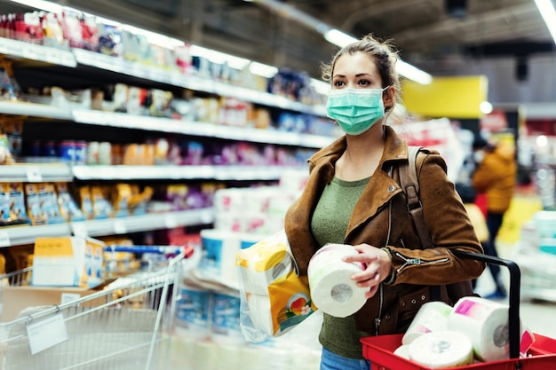 Jonge vrouw met beschermend masker die toiletpapier winkelt en benodigdheden maakt tijdens een viruspandemie