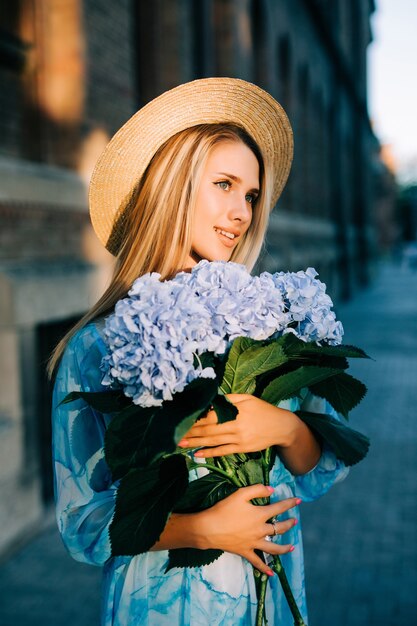 Jonge vrouw met blauwe hedraga buquet in blauwe jurk staande in groene tuin op zonsondergang