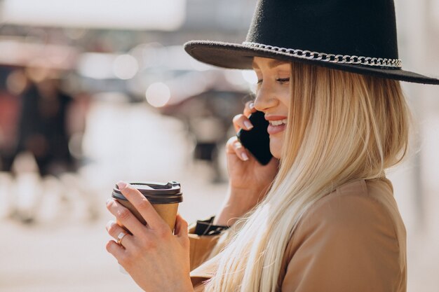 Jonge vrouw met blond haar die zwarte hoed draagt die aan de telefoon spreekt en koffie drinkt