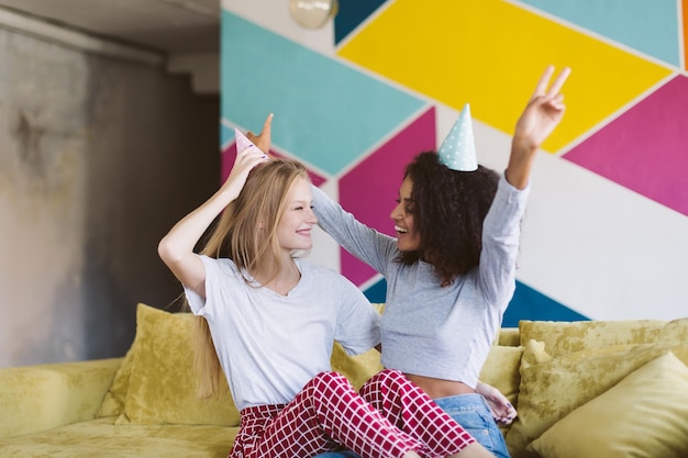 Jonge vrouw met blond haar en mooie Afro-Amerikaanse vrouw met donker krullend haar in verjaardagskappen die vreugdevol naar elkaar kijken terwijl ze tijd doorbrengen samen met kleurrijke muur op achtergrond