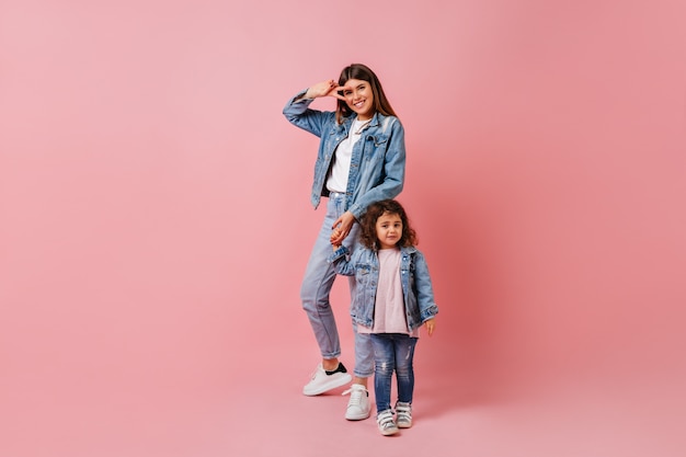 Jonge vrouw met dochter die vredesteken toont. Studio shot van geweldige stijlvolle dame hand in hand met kind op roze achtergrond.
