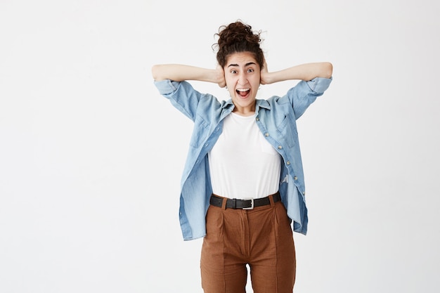 Gratis foto jonge vrouw met donker haar in broodje dat in verschrikking met handen op haar hoofd, mond wijd open het kijken in paniek gilt. geïrriteerd vrouwelijk geschreeuw, dat haar oren bedekt