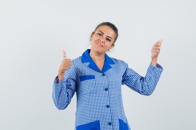 Jonge vrouw met dubbele duimen omhoog in het blauwe overhemd van de gingangpyjama en ziet er mooi uit. vooraanzicht.