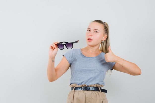 Jonge vrouw met duim omhoog, bril in t-shirt, broek vooraanzicht te houden.