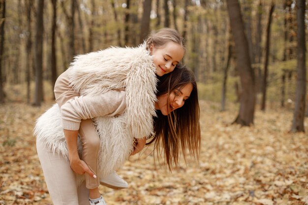 Jonge vrouw met een klein meisje dat zich in de herfstbos bevindt. Donkerbruine vrouw die haar dochter op de rug houdt. Meisje draagt beige trui en moeder draagt witte kleren.