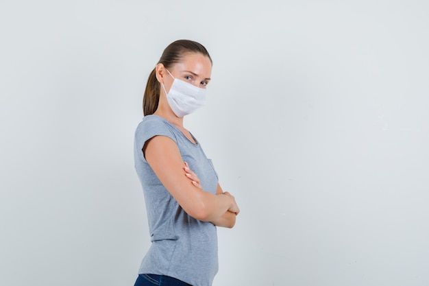 Jonge vrouw met gekruiste armen in t-shirt, masker, spijkerbroek en op zoek vrolijk.