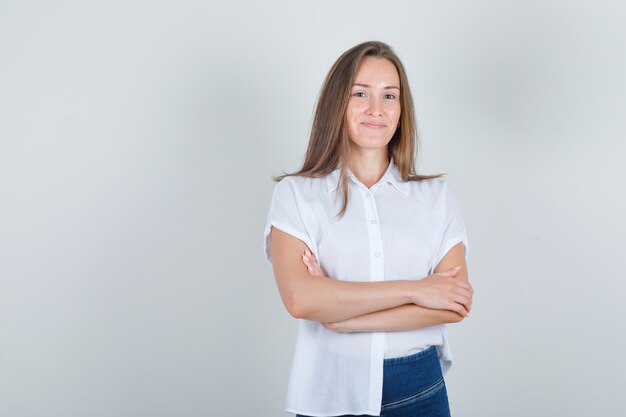 Jonge vrouw met gekruiste armen in wit t-shirt, spijkerbroek en op zoek vrolijk