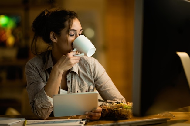 Jonge vrouw met gesloten ogen genietend van een kopje koffie terwijl ze thuis laat in touchpad werkt