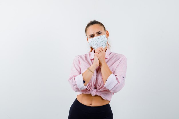 Jonge vrouw met gevouwen handen in smekend gebaar in shirt, broek, medisch masker en op zoek naar hoopvol, vooraanzicht.