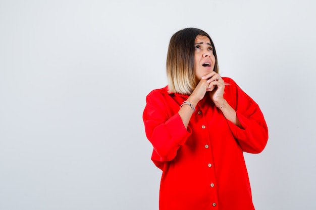 Jonge vrouw met gevouwen handen onder de kin in een rood oversized shirt en kijkt verbaasd, vooraanzicht.