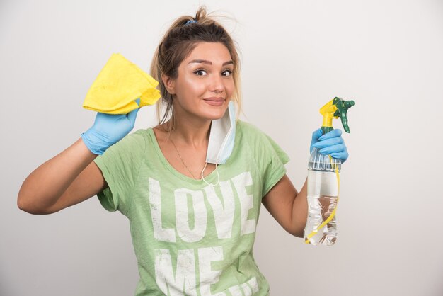 Jonge vrouw met gezichtsmasker en levert schoonmaakmiddelen op witte muur.