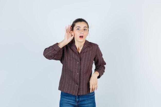 Jonge vrouw met hand in de buurt van oor om iets te horen, mond open houden in gestreept shirt, spijkerbroek en geschokt kijken, vooraanzicht.