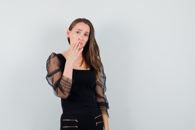 Jonge vrouw met hand op haar mond in zwarte blouse en op zoek bedroefd. vooraanzicht.