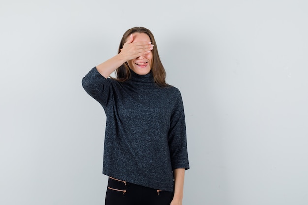 Jonge vrouw met hand op haar ogen terwijl tong uitsteekt in zwarte blouse en verlegen kijkt