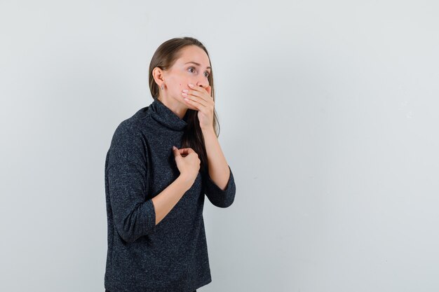 Jonge vrouw met hand op mond in shirt en op zoek angstig. vooraanzicht.