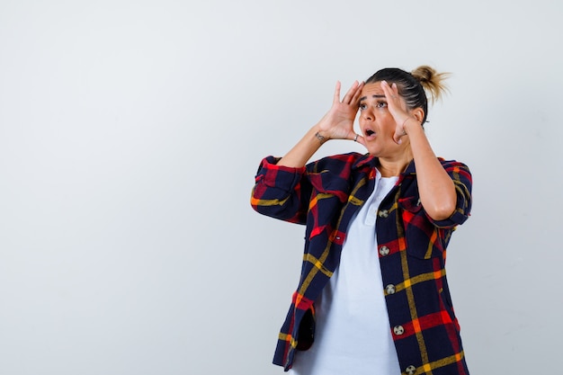 Jonge vrouw met handen om duidelijk te zien, zijwaarts in geruit overhemd en geschokt kijkend.