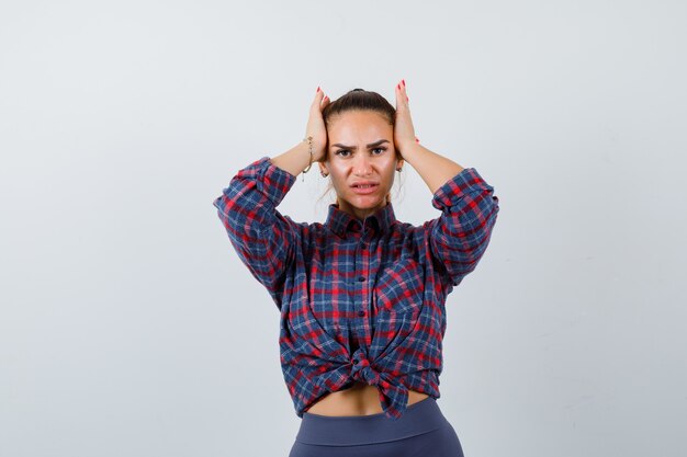 Jonge vrouw met handen op het hoofd in geruit overhemd, broek en vergeetachtig kijken. vooraanzicht.