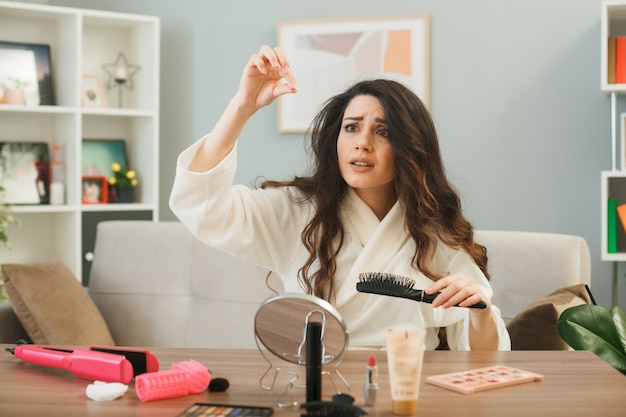 Jonge vrouw met kam zittend aan tafel met make-up tools in woonkamer