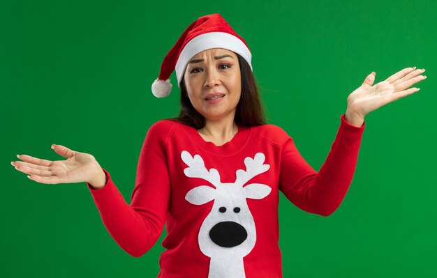 Jonge vrouw met kerst kerstmuts en rode trui op zoek verward schouderophalend staande over groene achtergrond