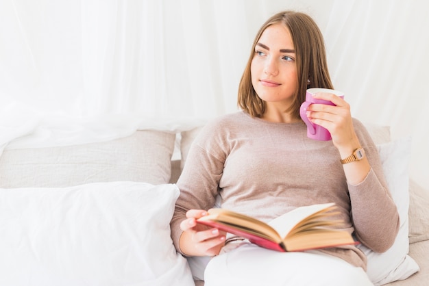 Gratis foto jonge vrouw met koffie zittend op bed bedrijf boek