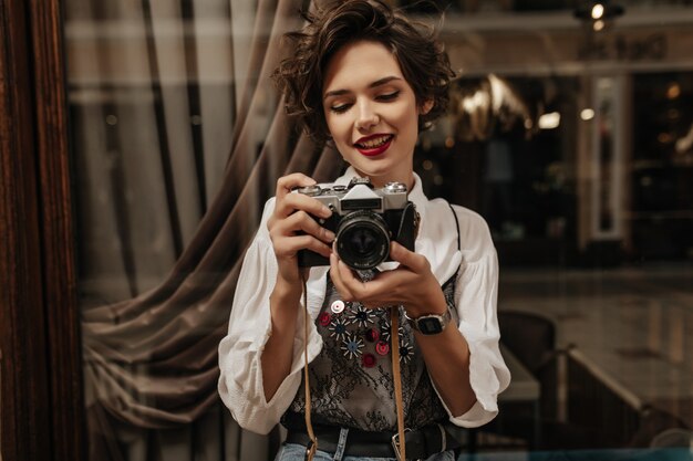 Jonge vrouw met kort haar en rode lippenstift die foto binnen maken. Positieve vrouw in een stijlvol wit overhemd met camera in café.