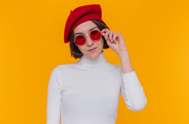 Jonge vrouw met kort haar in een witte coltrui die een baret en een rode zonnebril draagt en naar de voorkant kijkt glimlachend zelfverzekerd blij en positief staande over oranje muur