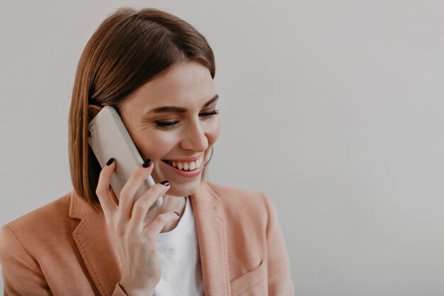 Jonge vrouw met kort haar in fel jasje glimlacht en spreekt via de telefoon op wit.