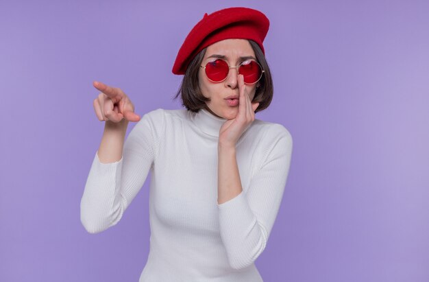 Jonge vrouw met kort haar in witte coltrui met baret en rode zonnebril op zoek verrast en geïntrigeerd met wijsvinger wijzend naar iets dat over blauwe muur staat