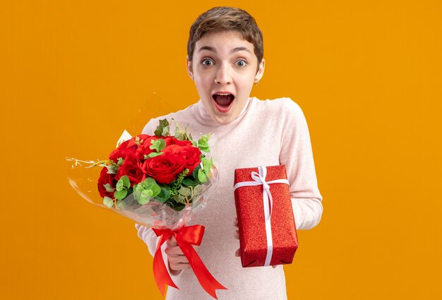 jonge vrouw met kort haar met boeket van rode rozen en een cadeau kijkend naar de camera verbaasd en verrast Valentijnsdag concept staande over oranje muur