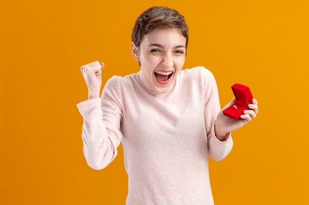 jonge vrouw met kort haar met rode doos met verlovingsring blij en opgewonden gebalde vuist blij en opgewonden Valentijnsdag concept staande over oranje muur