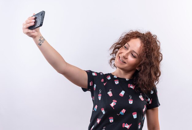 Jonge vrouw met kort krullend haar die selfie met haar smartphone gebruiken die aan camera glimlacht die zich over witte muur bevindt