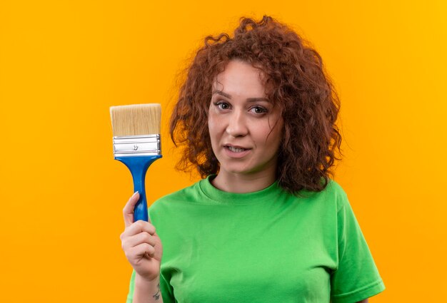 Jonge vrouw met kort krullend haar in de groene kwast van de t-shirtholding die zelfverzekerd status kijkt