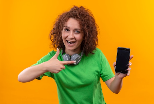 Jonge vrouw met kort krullend haar in groen t-shirt die smartphone toont die met vinger ernaar richt glimlachend breed staande over oranje muur
