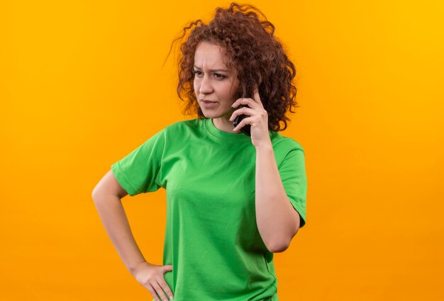 Jonge vrouw met kort krullend haar in groen t-shirt op zoek verward en erg angstig tijdens het praten over de mobiele telefoon