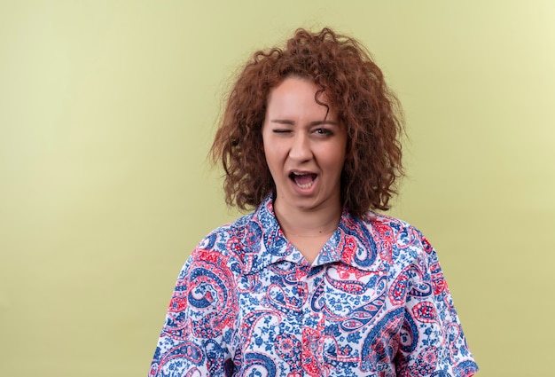 Jonge vrouw met kort krullend haar in kleurrijk overhemd knipogen en geeuw kijkt moe over groene muur