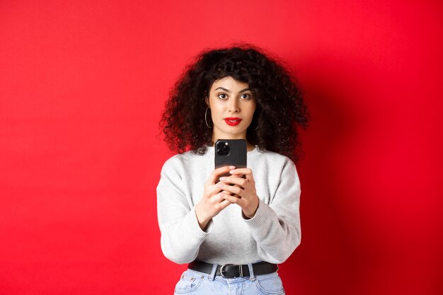 Jonge vrouw met krullend haar, video opnemen op smartphone, foto nemen op mobiele telefoon en camera kijken, staande op rode achtergrond.