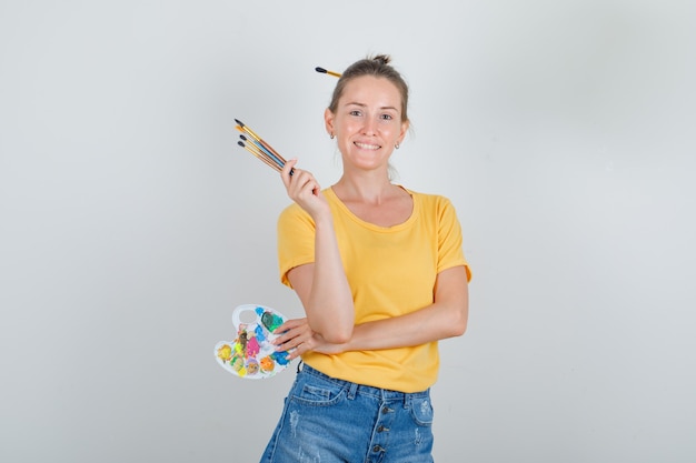 Jonge vrouw met kunst palet met borstels in geel t-shirt, jeans broek en op zoek gelukkig