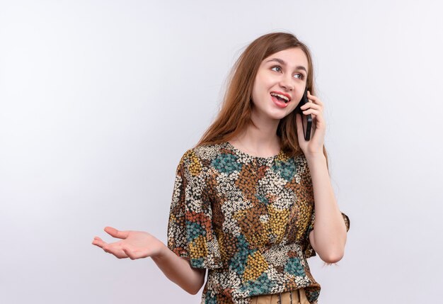 Jonge vrouw met lang haar die kleurrijke kleding dragen die tijdens het spreken op mobiele telefoon over witte muur glimlacht