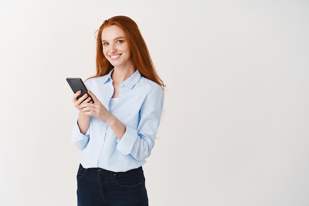 Jonge vrouw met lang rood haar en witte glimlach die gelukkig naar voren kijkt, met behulp van mobiele telefoon, witte muur