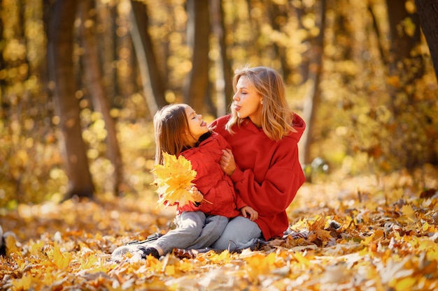 Jonge vrouw met meisje zittend op een deken in herfst bos. Blonde vrouw speelt met haar dochter. Moeder en dochter dragen jeans en rode jassen.