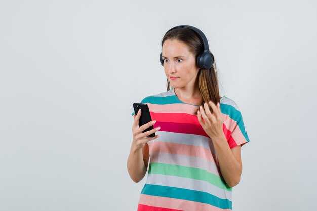 Jonge vrouw met mobiele telefoon in t-shirt, koptelefoon en boos kijkt. vooraanzicht.