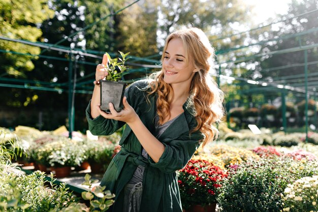 Jonge vrouw met mooi blond haar en zachte glimlach, gekleed in groen gewaad met riem werkt in kas