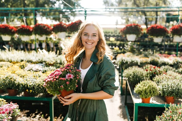 Jonge vrouw met mooi blond haar en zachte glimlach, gekleed in groen gewaad met riem werkt in kas