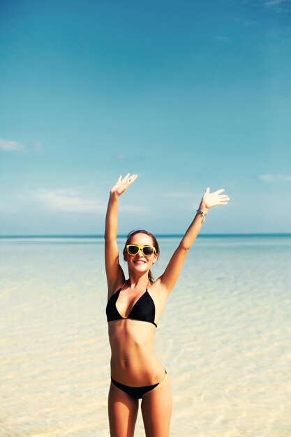 Jonge vrouw met opgeheven handen en gele zonnebril op het strand